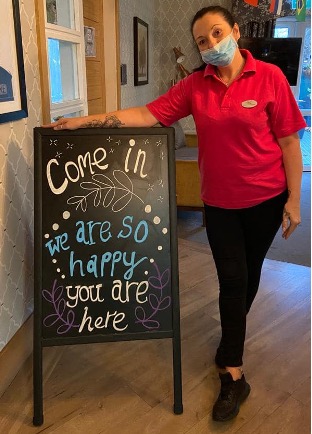 A member of staff next to a sign reading "Come in we are so happy you are here" at Haling Park Care Home